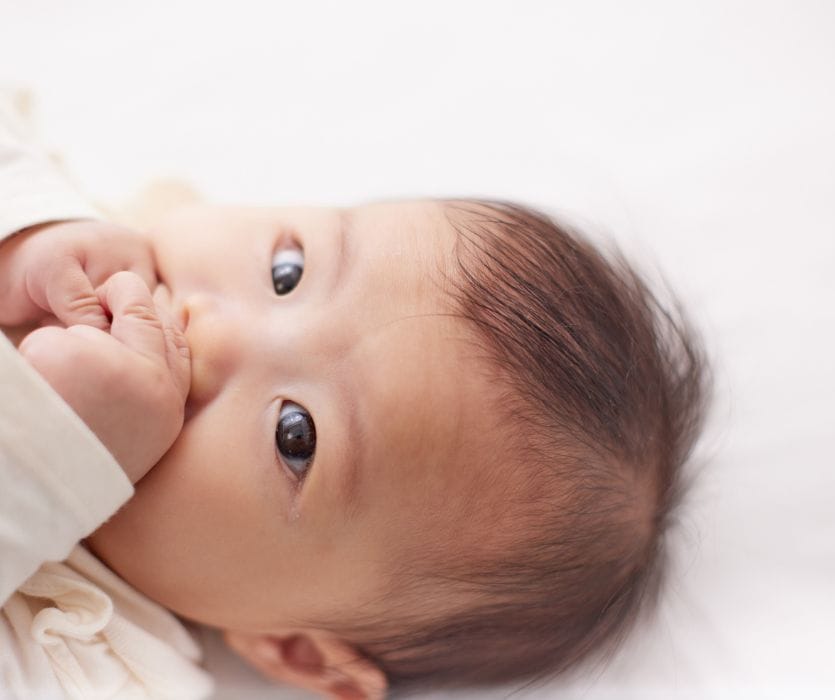 A baby lying on their back with their hand in their mouth, looking toward the camera.