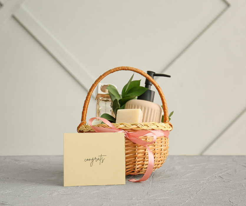 Wicker gift basket with a pink bow, toiletries, green leaves, and a congrats card on a grey surface.