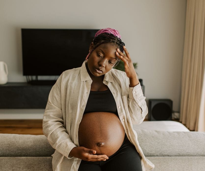 A pregnant woman sitting on a sofa, holding her head in pain and touching her belly.