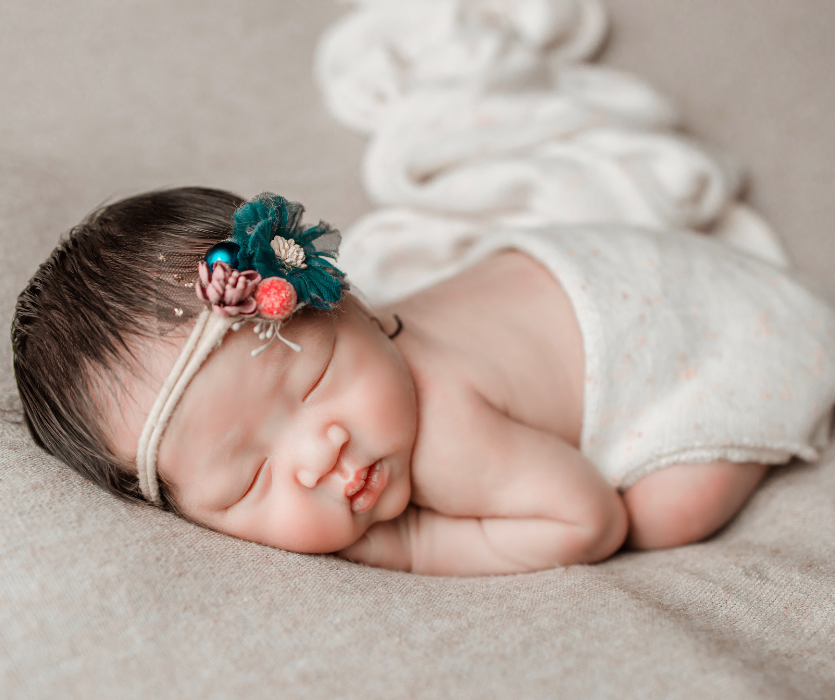 Sleeping newborn with dark hair wearing a floral headband and white wrap on a tan blanket.
