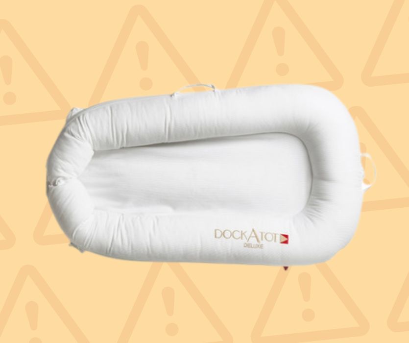 White DockATot Deluxe baby lounger on a yellow background with triangle warning icons.