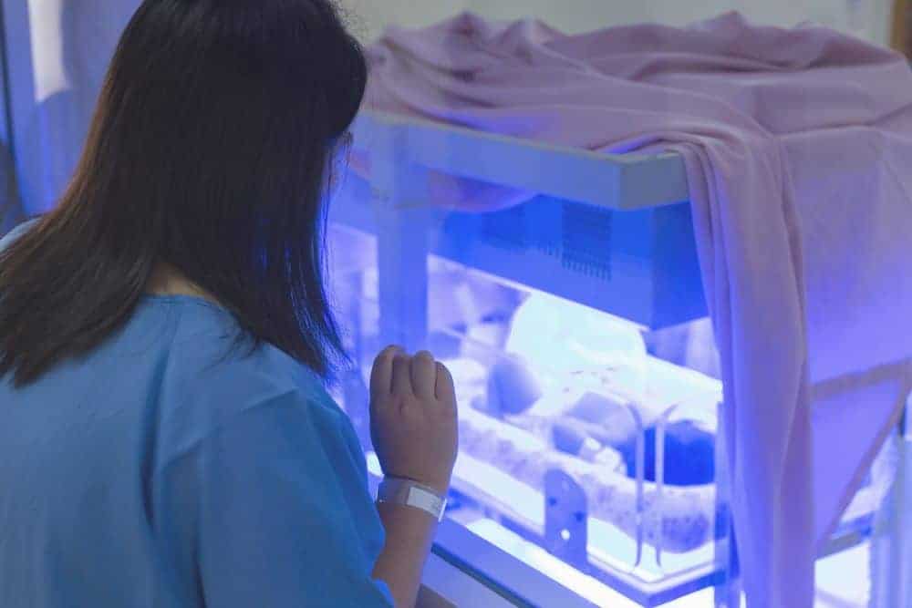 A woman in a hospital gown looks into a NICU incubator where a newborn receives blue light phototherapy for jaundice.