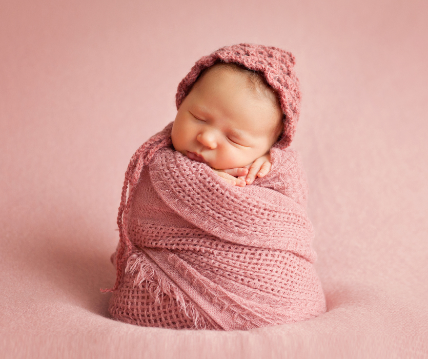 Sleeping newborn baby swaddled in a pink knit wrap and matching bonnet against a pink background.