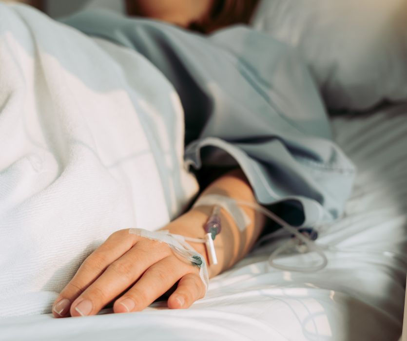 A person lying in a hospital bed with an IV line inserted into the back of their hand.