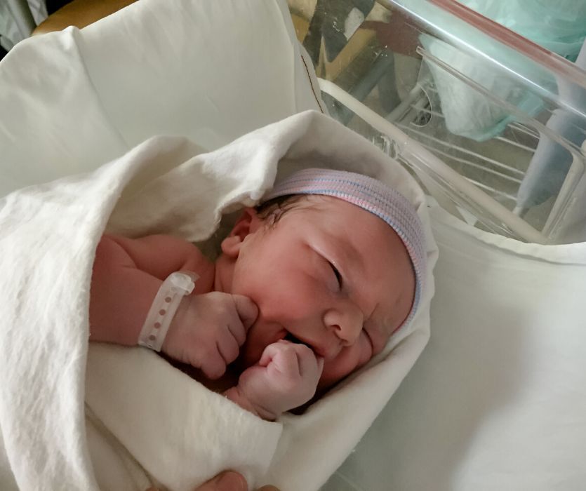 Swaddled newborn baby wearing a pink and blue striped hospital hat and ID bracelet, resting in a white bassinet.