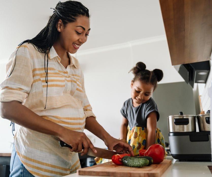 Smiling pregnant woman slicing vegetables on a wooden board next to a young girl in a kitchen.