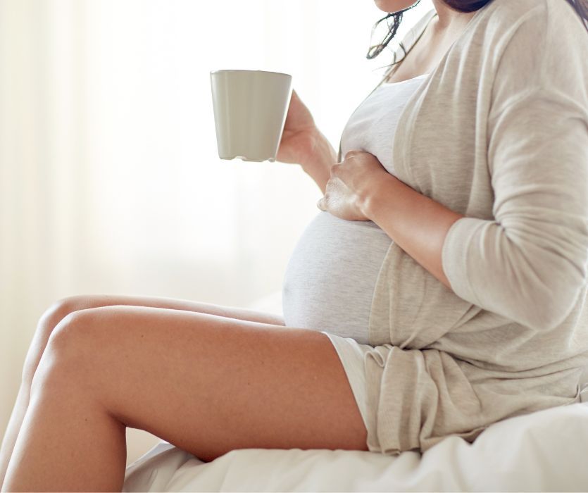 Pregnant woman sitting on a bed, holding a mug and resting her hand on her belly.