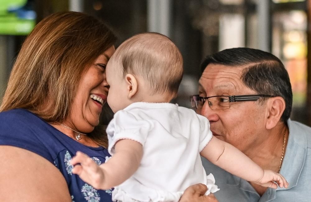 A smiling woman and a man in glasses lean in toward a baby held in the foreground.