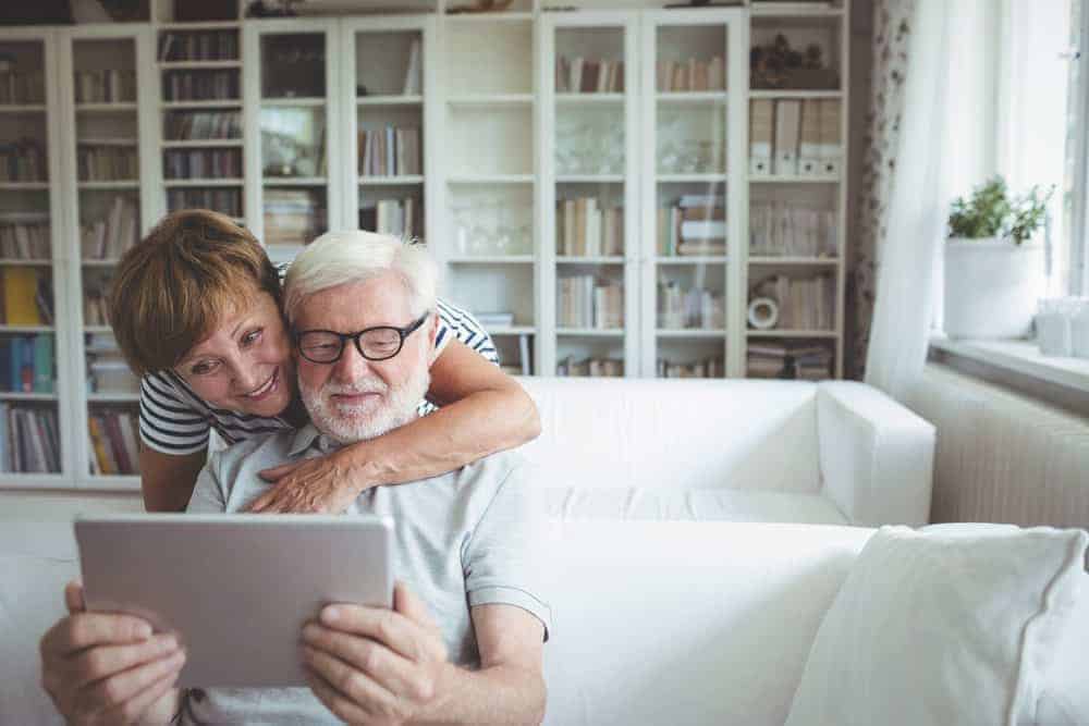A smiling senior couple uses a digital tablet while sitting on a white sofa in a living room with bookshelves.