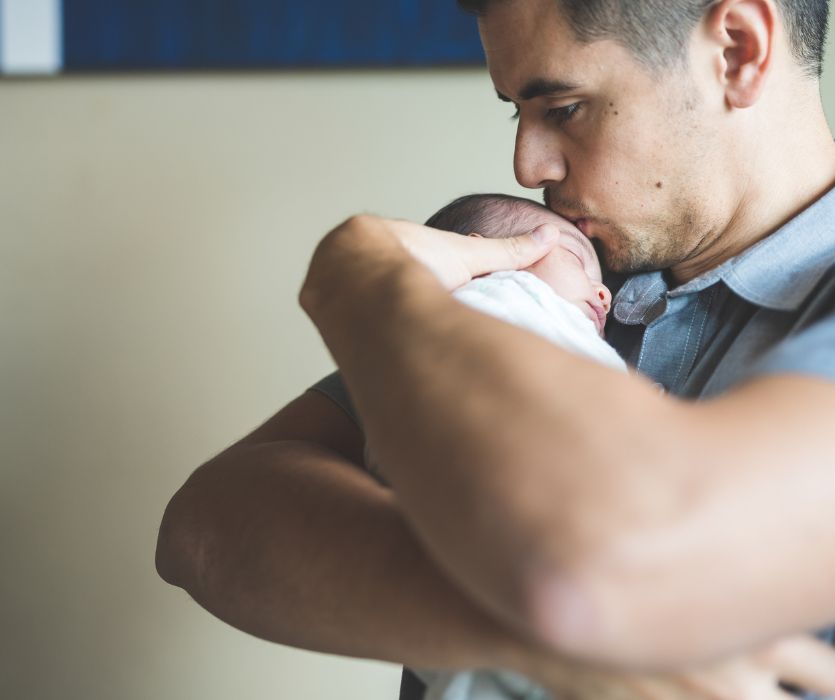 A man cradles a sleeping newborn baby in his arms and kisses the infant's forehead.