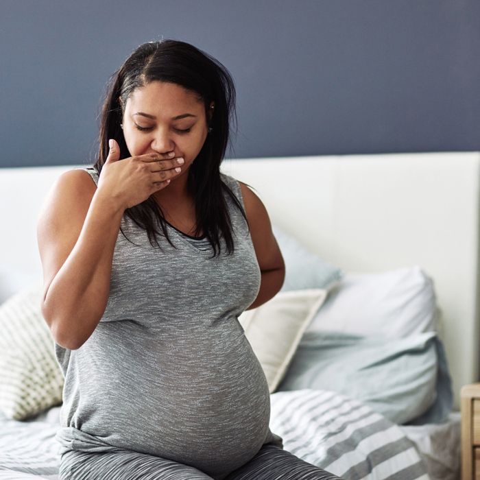 Pregnant woman sitting on a bed, holding her hand over her mouth and clutching her back in discomfort.