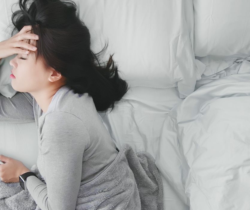 A woman lying in bed with her hand on her forehead, looking distressed or unwell.