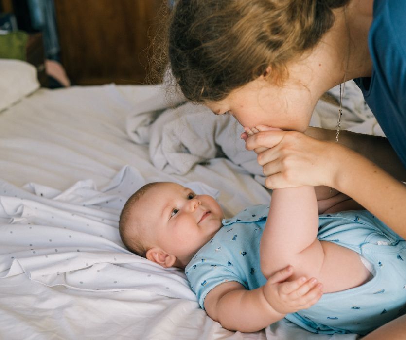 A mother kisses the foot of her smiling baby lying on a bed.