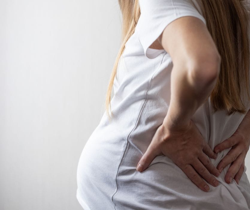 Side view of a pregnant person in a white shirt holding their lower back in discomfort.