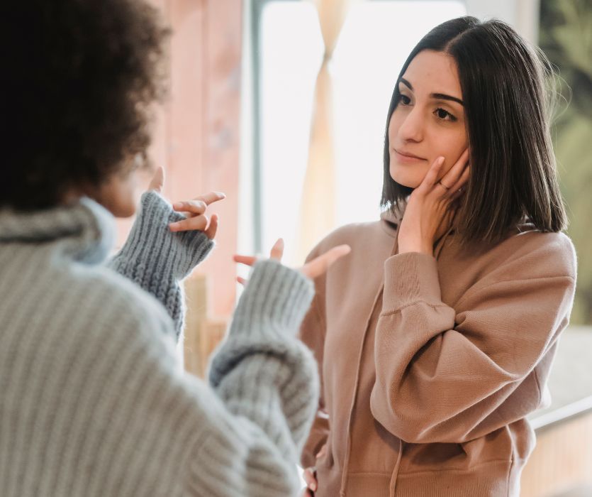 A woman in a brown hoodie listens with a concerned expression as another person gestures while talking to her.
