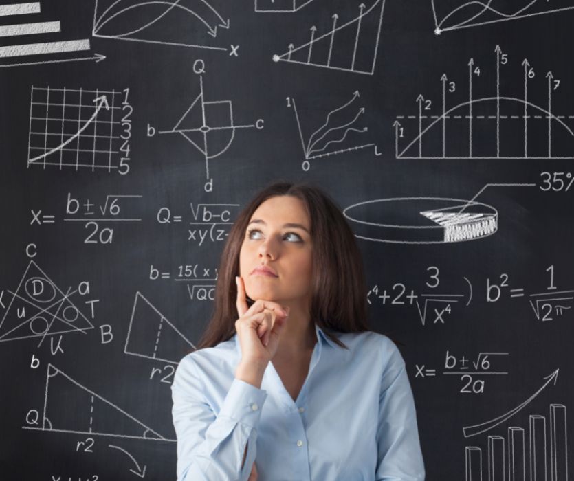 Thinking woman in a blue shirt standing in front of a chalkboard covered in complex math equations and diagrams.