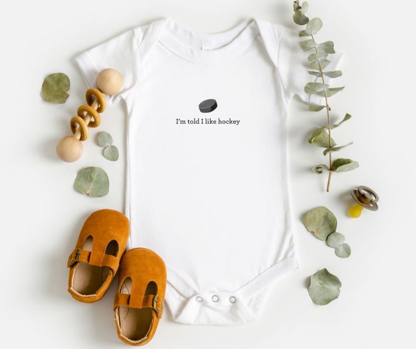 White baby bodysuit with a hockey puck graphic and the text I'm told I like hockey, surrounded by baby shoes and toys.