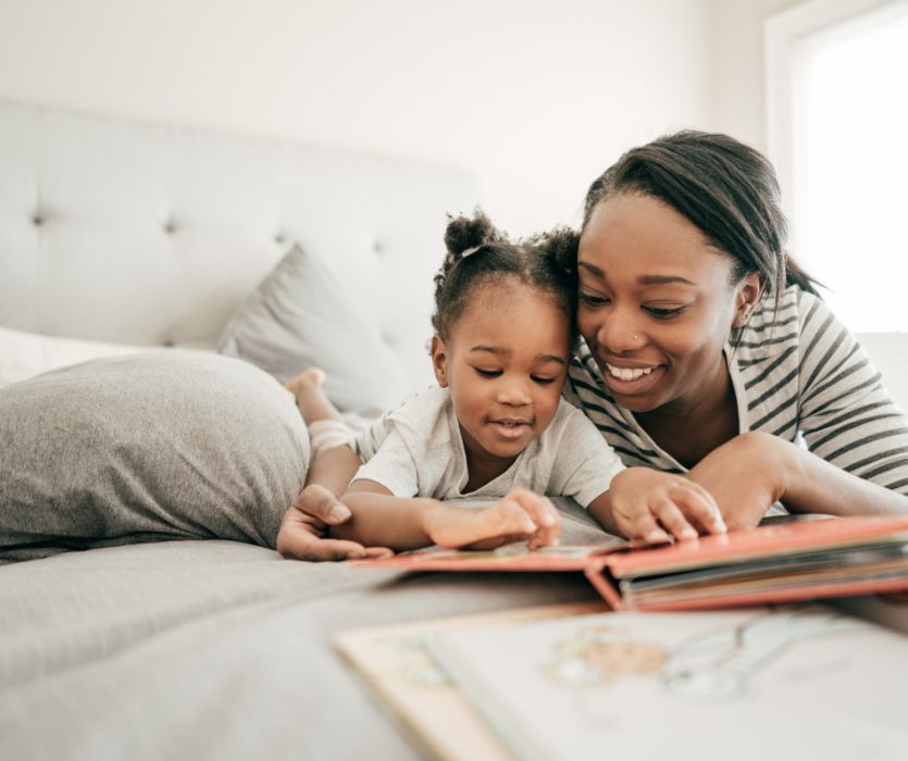 A smiling Black woman and a young girl lying on a bed reading a picture book together.