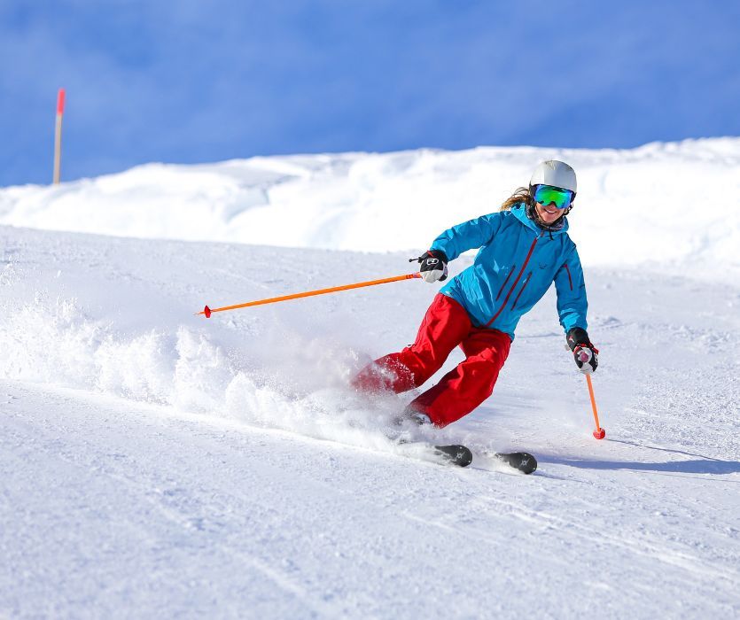 A smiling woman in a blue jacket and red pants skis down a snowy mountain slope under a blue sky.
