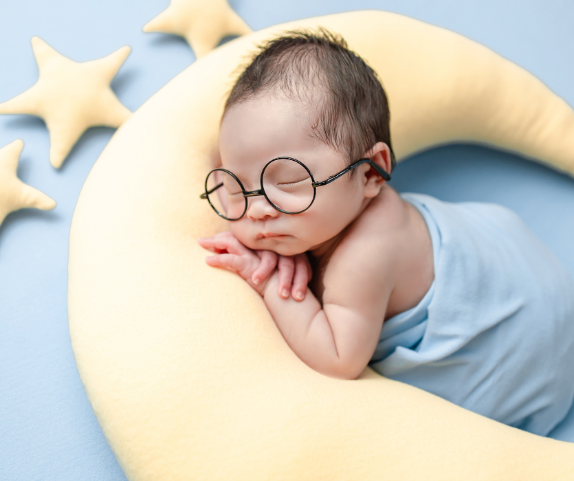 A newborn baby wearing round glasses, sleeping on a yellow crescent moon-shaped pillow against a blue background.