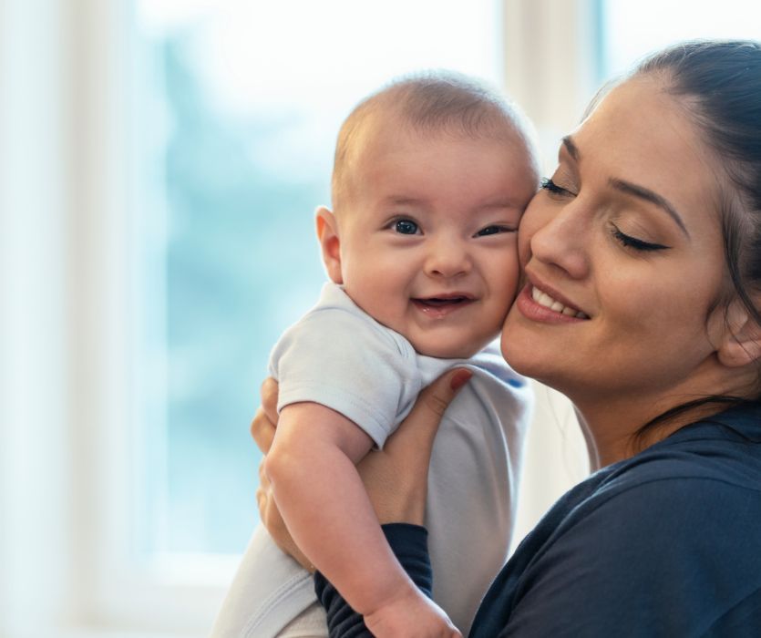 A smiling woman holds a happy infant close to her cheek against a blurred window background.