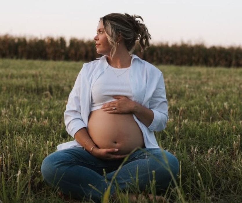 Laurie Bagwell sits in a grassy field, cradling her bare pregnant belly while looking to the side.