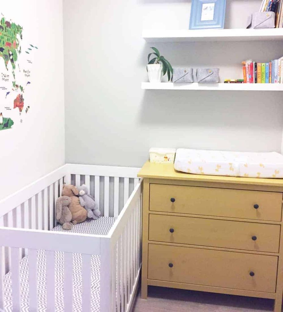 White crib with stuffed animals next to a wooden dresser with a changing pad and wall shelves with books and plants.