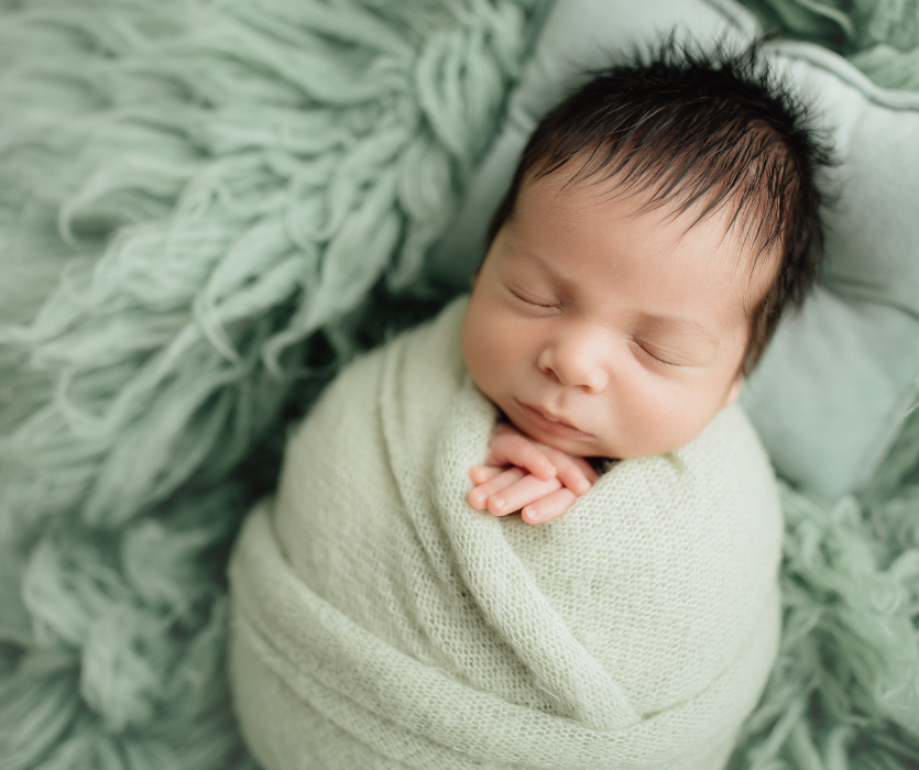 A sleeping newborn with dark hair, wrapped in a light green swaddle, resting on a textured green blanket.