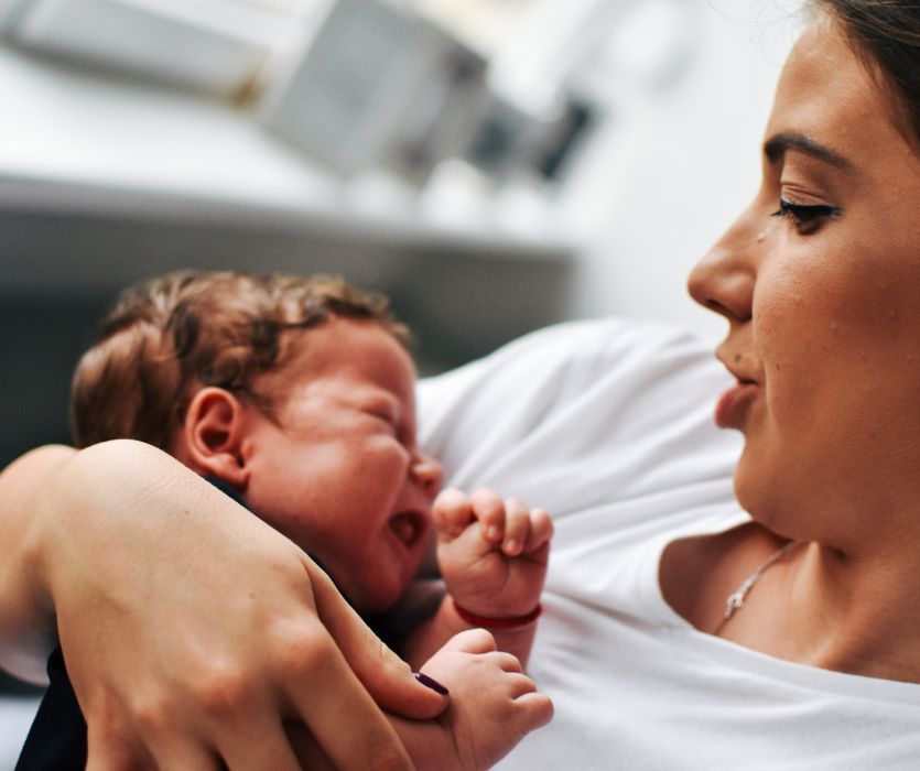 A woman in a white t-shirt looks down lovingly at a crying newborn baby cradled in her arms.