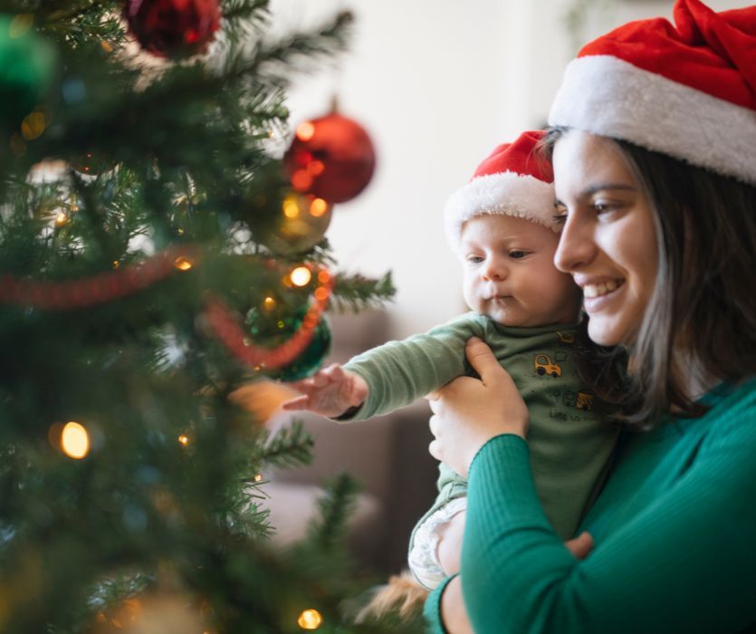 A woman and baby wearing Santa hats smile while looking at a decorated Christmas tree.