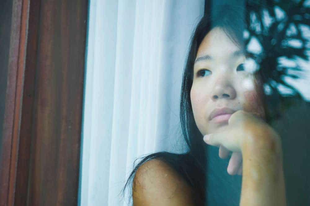 A woman looks out a window with a somber, pensive expression, resting her chin on her hand.