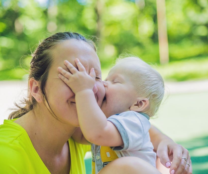 A smiling woman in a yellow shirt cuddles a toddler who is kissing her cheek and covering her eyes with his hand.