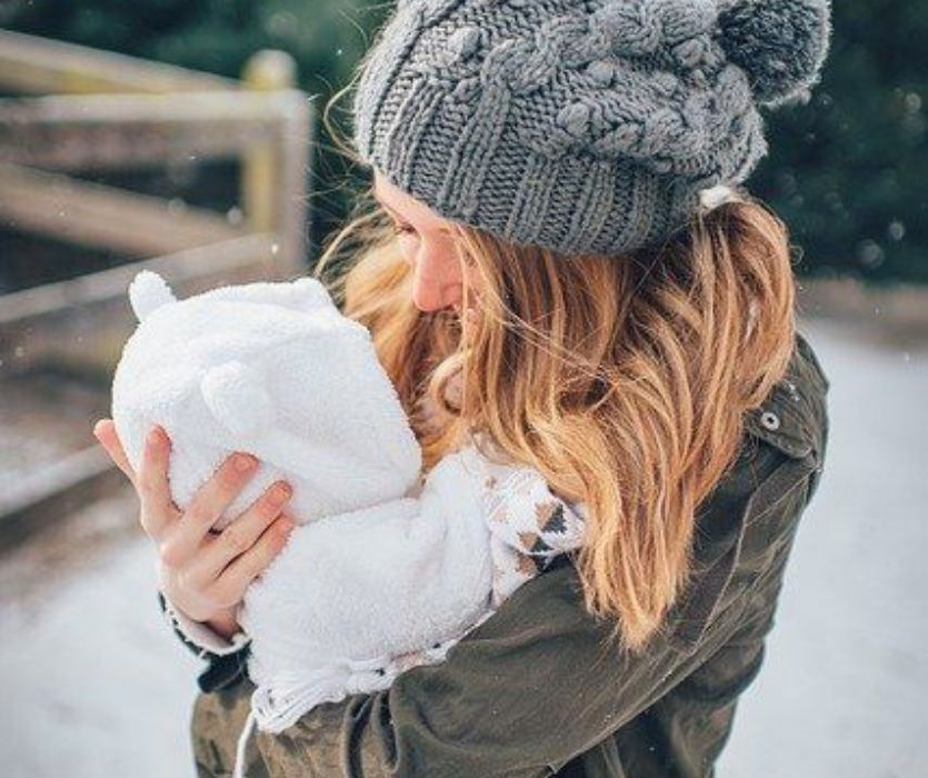 A woman in a gray knit beanie holds a baby wearing a white fuzzy snowsuit with animal ears outside in the snow.