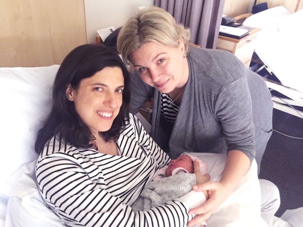 A woman smiling in a hospital bed holding a newborn baby next to another woman wearing a grey cardigan.