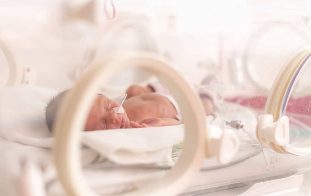 A newborn baby with medical tubing sleeping inside a high-tech hospital incubator in a NICU.