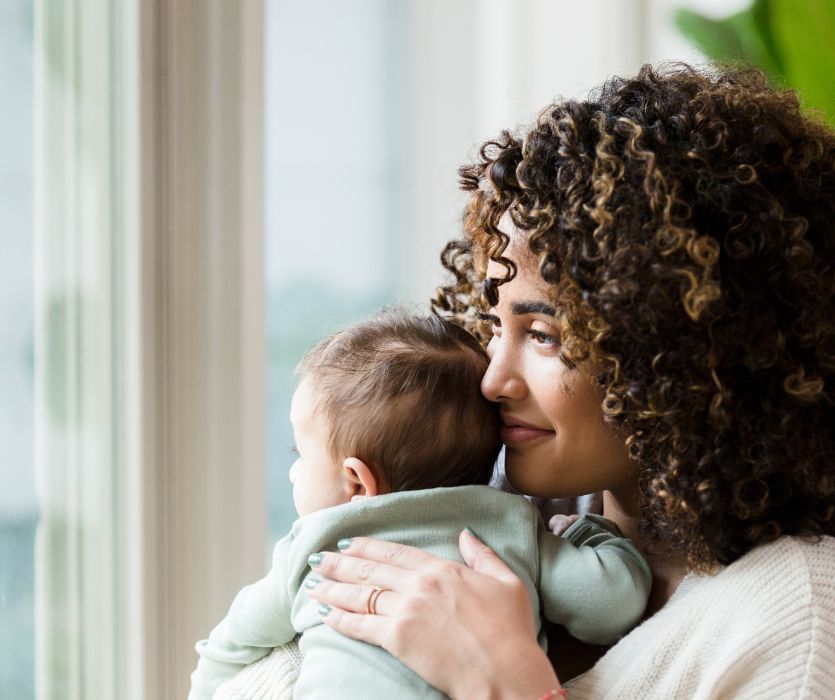 A woman with curly hair looks thoughtfully out a window while holding her infant.