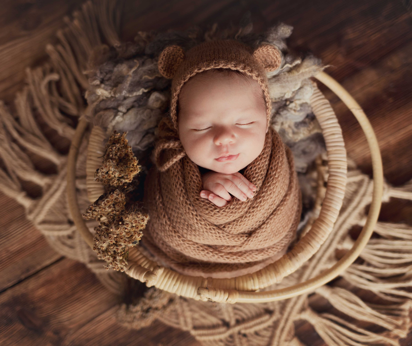 Sleeping newborn swaddled in a brown knit wrap and bear ear hat, resting in a woven basket on a wooden floor.