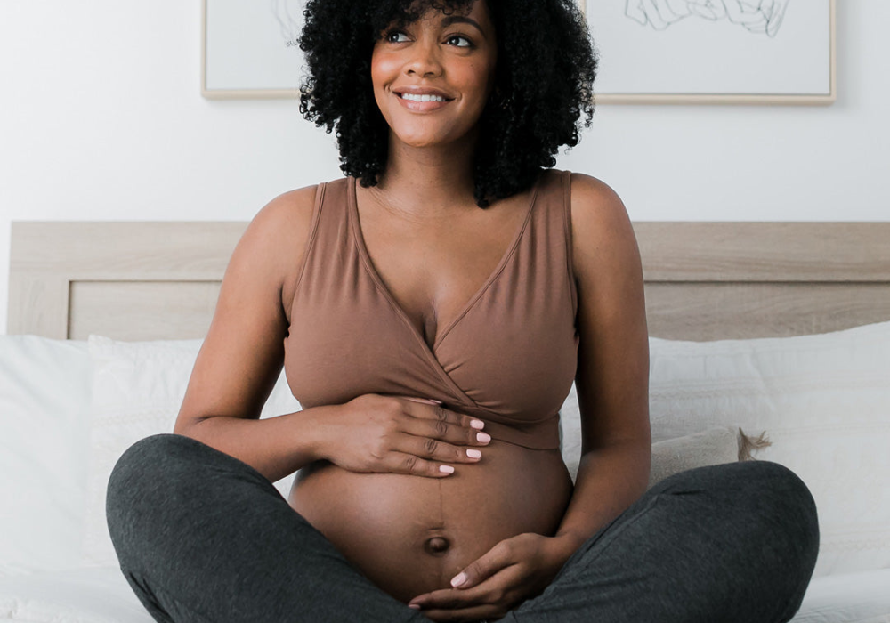 Pregnant person with curly hair smiling and holding their bare belly while wearing a brown crossover nursing bra.