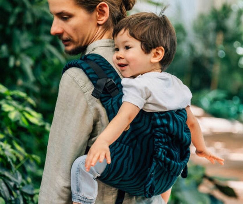 A smiling toddler in a white shirt sits in a teal-patterned onbuhimo baby carrier on a man's back.