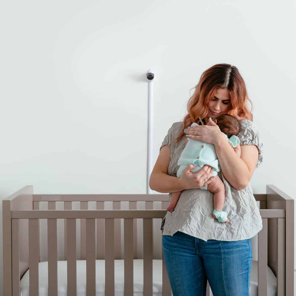 Woman holds a baby wearing an Owlet Smart Sock beside a crib with an Owlet Cam mounted on the wall behind them.