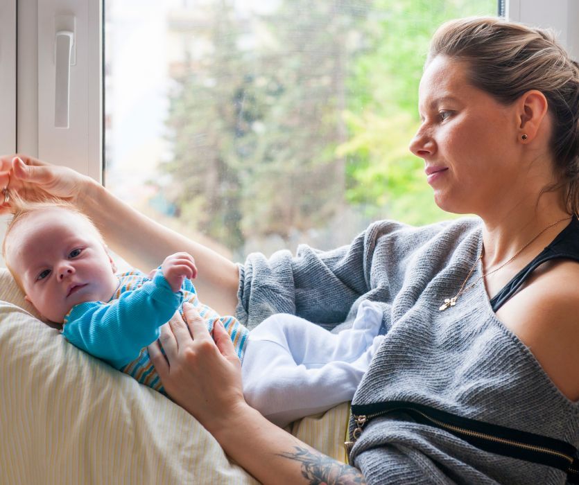 A woman sitting by a window cradles a baby in her lap while gently touching the infant's hair.