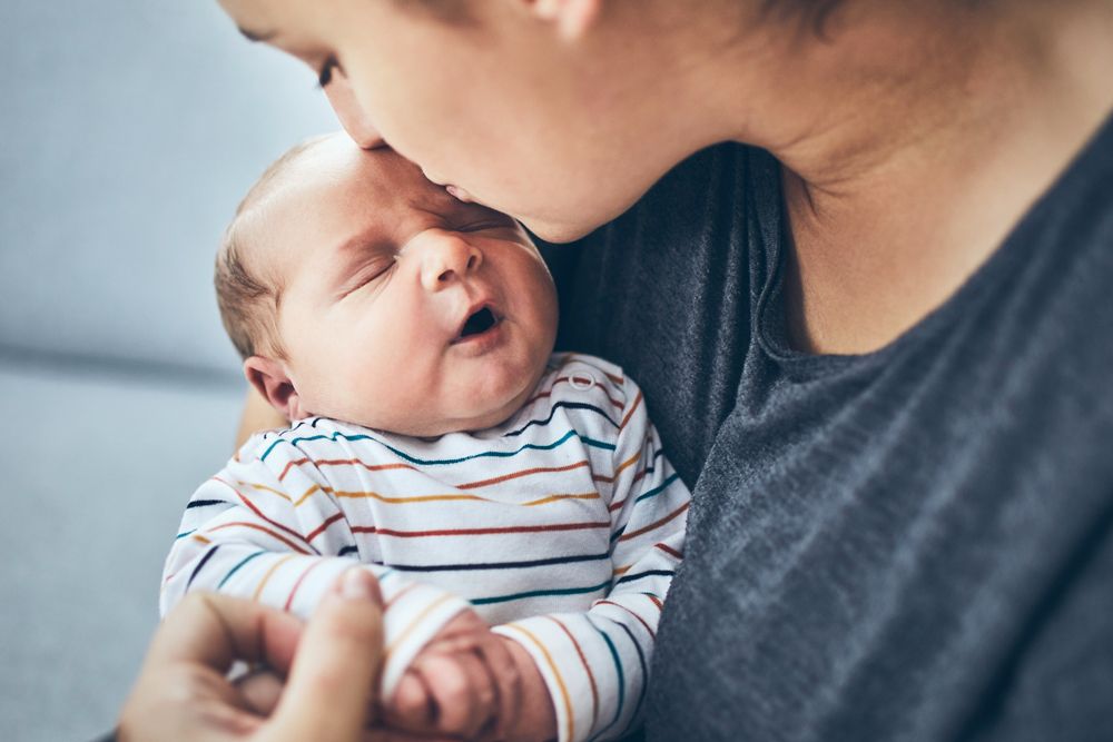 A woman kisses the forehead of a yawning newborn baby wearing a striped onesie.