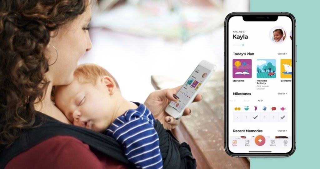 A woman holds a sleeping baby in a sling while using a parenting app on her phone to track daily plans and milestones.