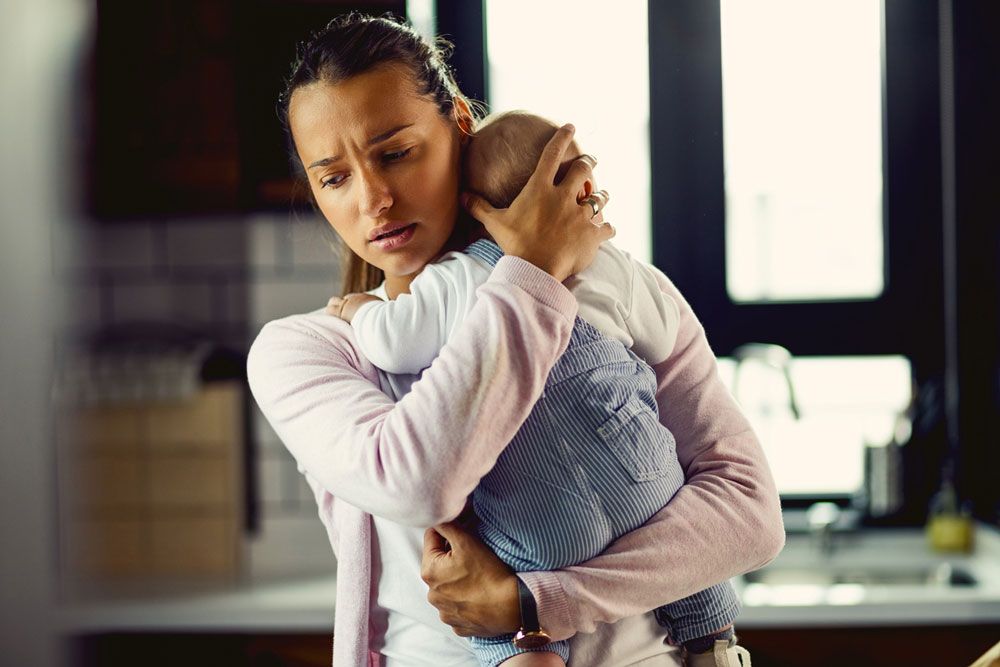 A distressed woman holds an infant closely while looking away with a worried, overwhelmed expression.
