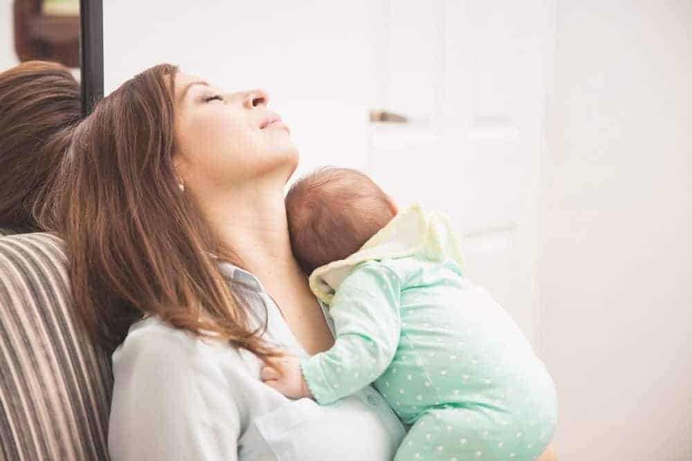 Tired mother with eyes closed holding a sleeping infant against her chest.