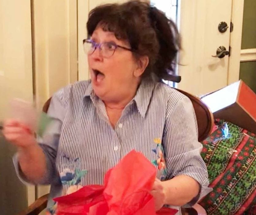 A woman with glasses and dark hair looks surprised with her mouth open as she opens a gift with red tissue paper.