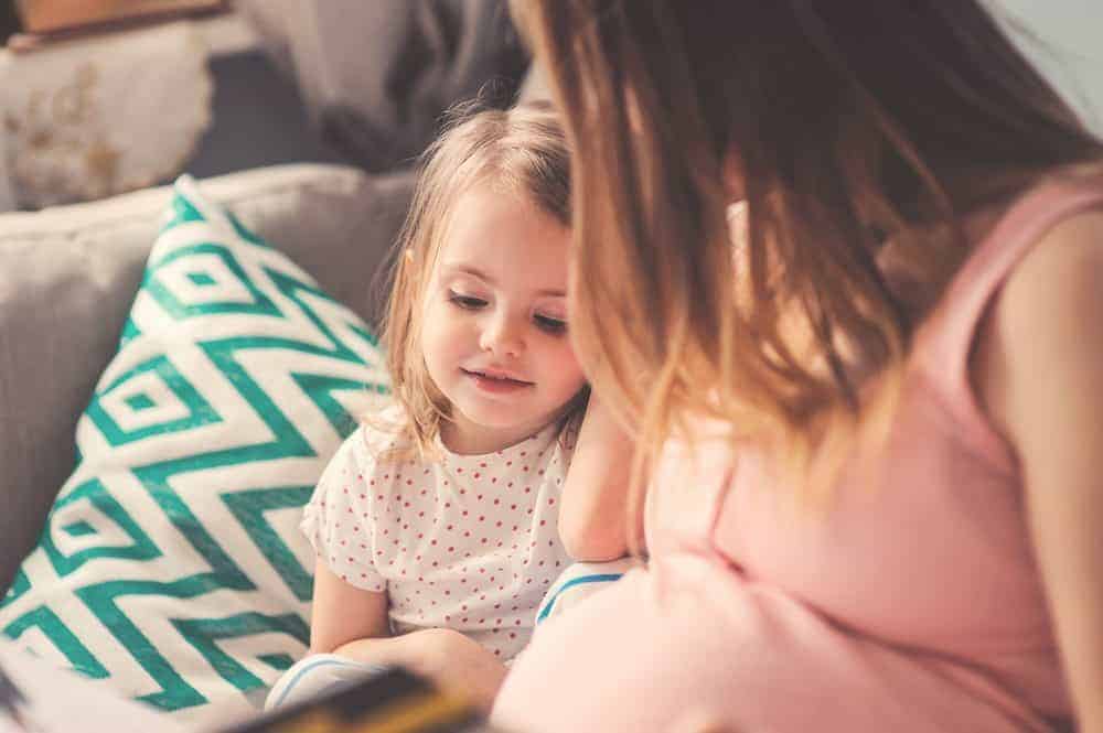 A young girl looking down toward her pregnant mother's stomach while sitting together on a couch.