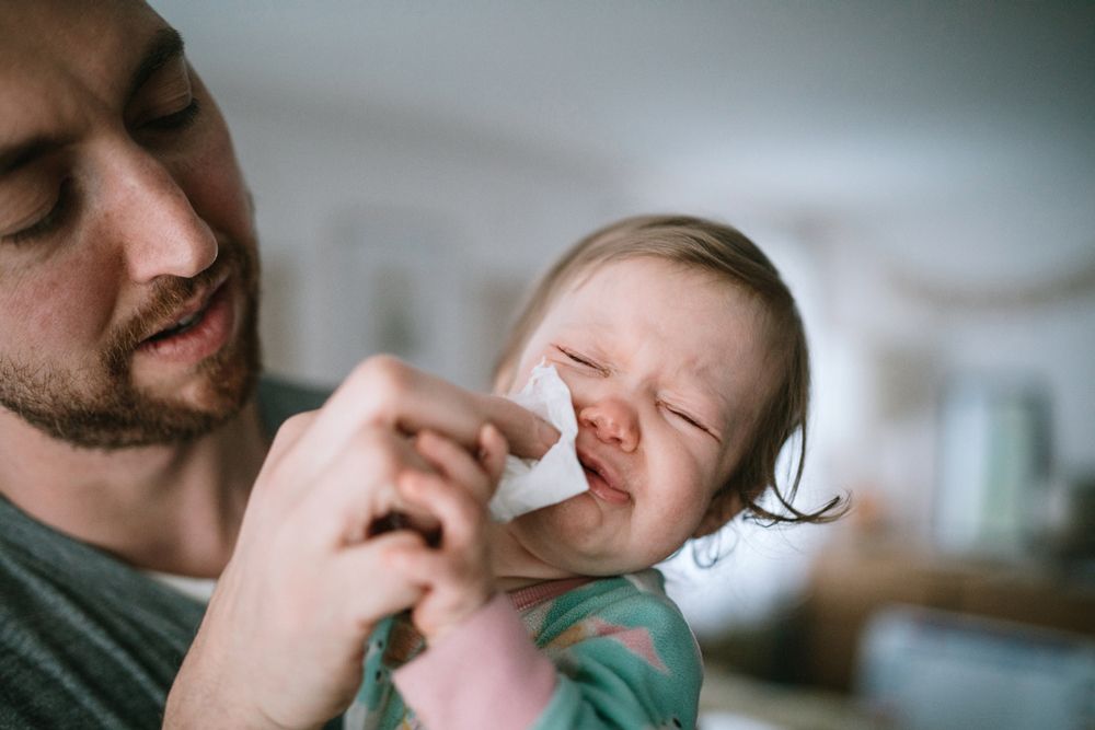A man uses a tissue to wipe the face of a crying toddler who has a runny nose.