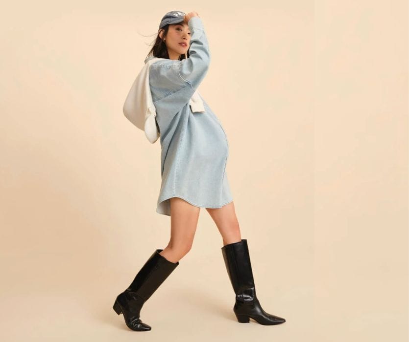 Pregnant woman in a denim shirt dress, black boots, and baseball cap, posing against a neutral background.