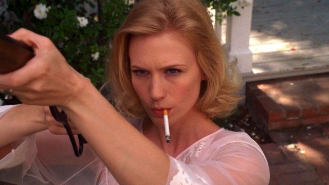 A woman with blonde hair smoking a cigarette while aiming a rifle outdoors.